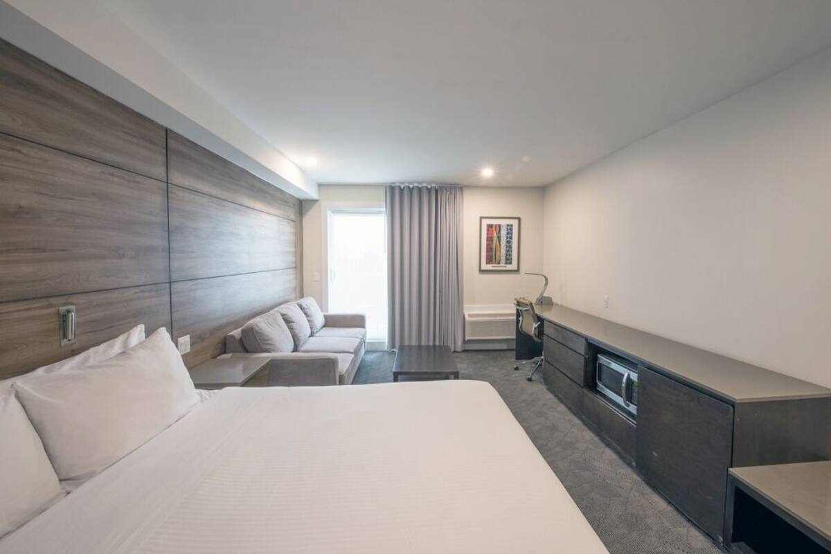 A modern hotel room with a large bed, a sofa, a desk, and a built-in oven.