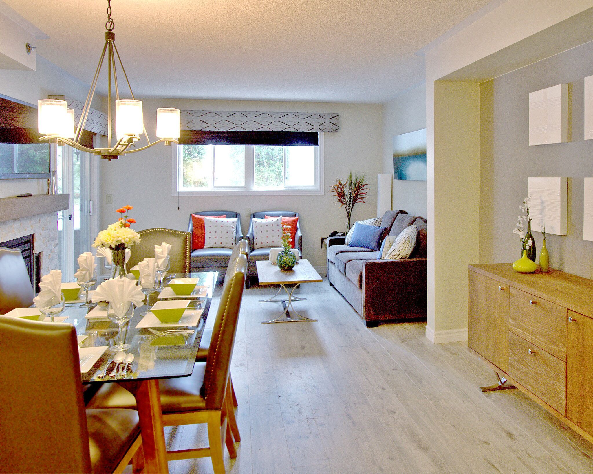 A dining area with a glass table, chairs, and a chandelier. There is a living room with a sofa, a coffee table, and a TV. The room has hardwood floors and a fireplace.