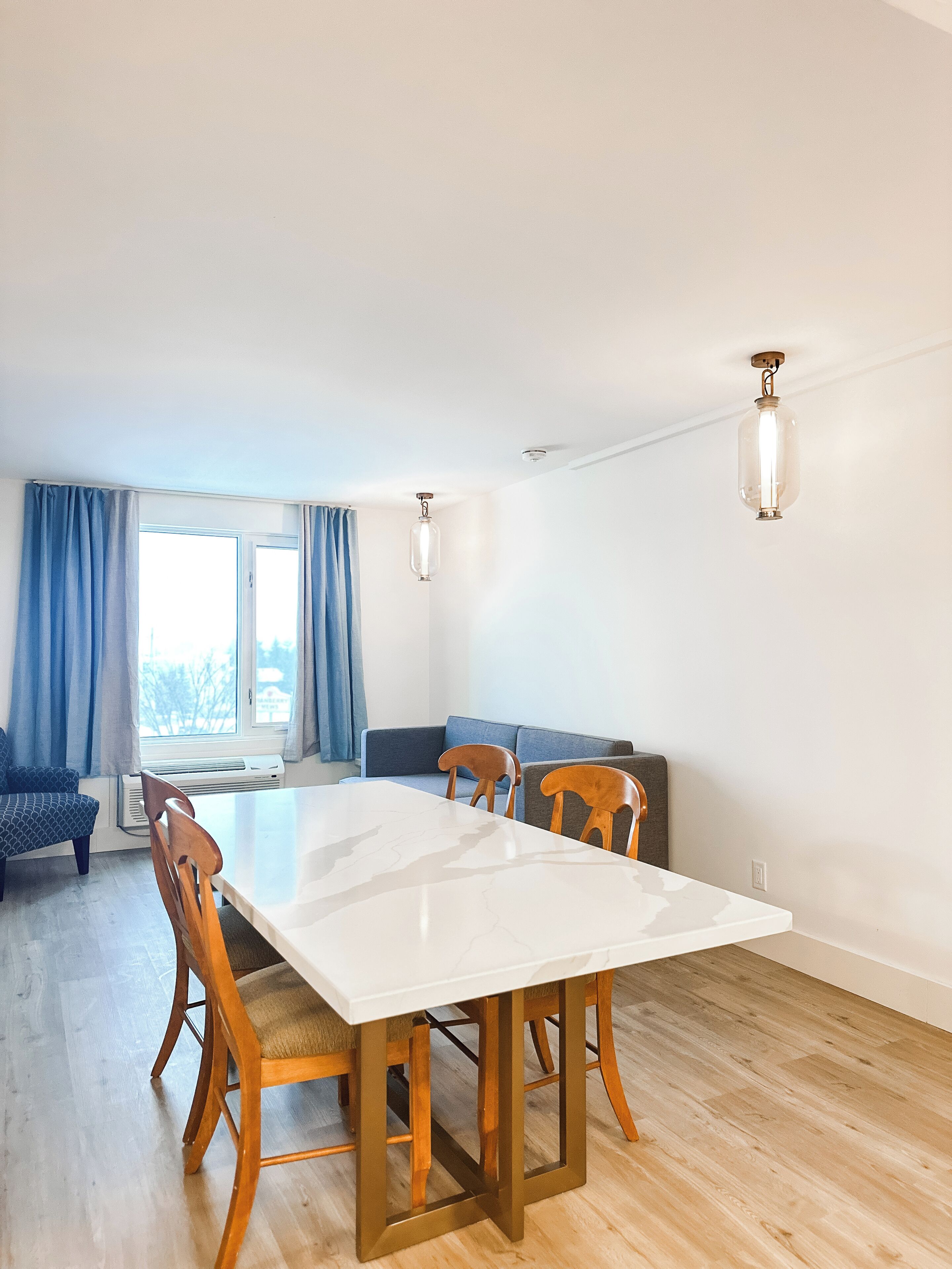 Zona pranzo con tavolo bianco, sedie di legno, divano blu e due lampadari.