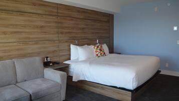 Ein modernes Hotelzimmer mit einem Holz-Kopfteil, einem Bett mit weißer Bettwäsche, einem grauen Sofa und einem kleinen Nachttisch.