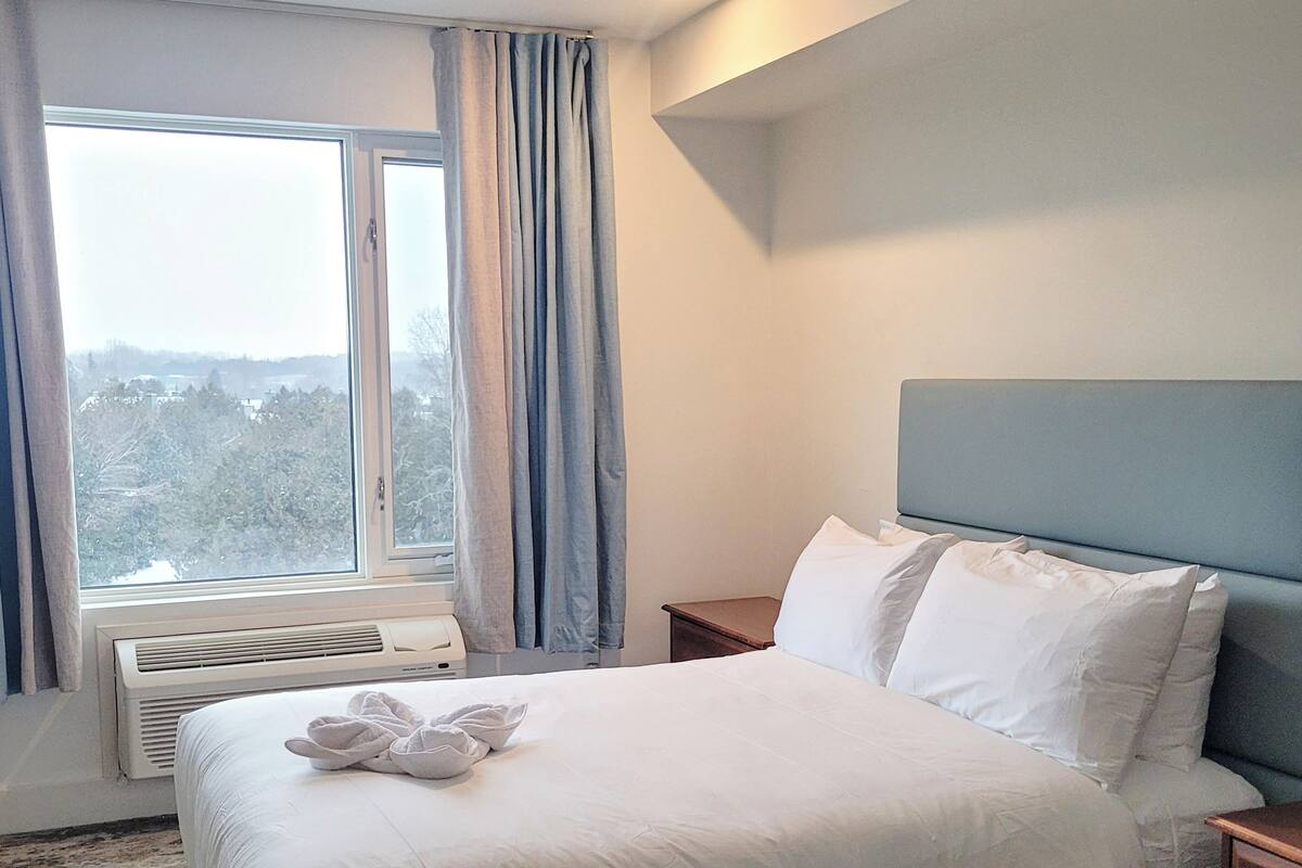 A neatly made bed with white linens and a towel, a bedside table, a nightstand, a window with curtains, and a wall-mounted air conditioner.