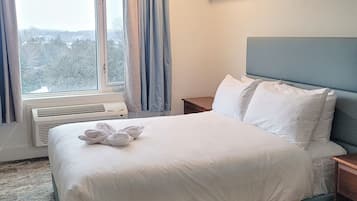 Una cama bien hecha con sábanas blancas y una toalla, una mesita de noche, una ventana con cortinas y un aire acondicionado de pared.