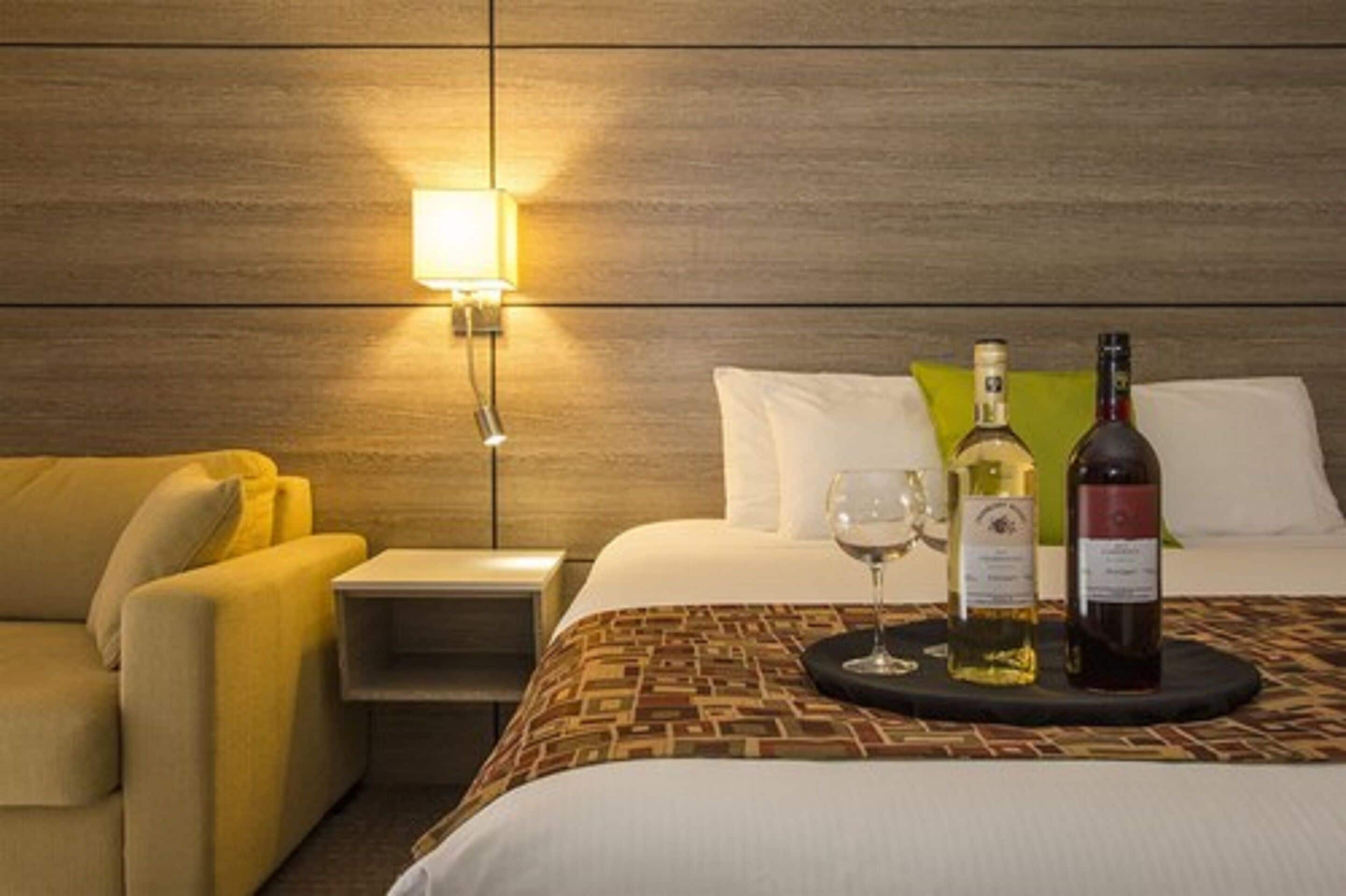 Une chambre d’hôtel avec un lit, une table de chevet, une lampe, un canapé et deux bouteilles de vin sur un plateau.