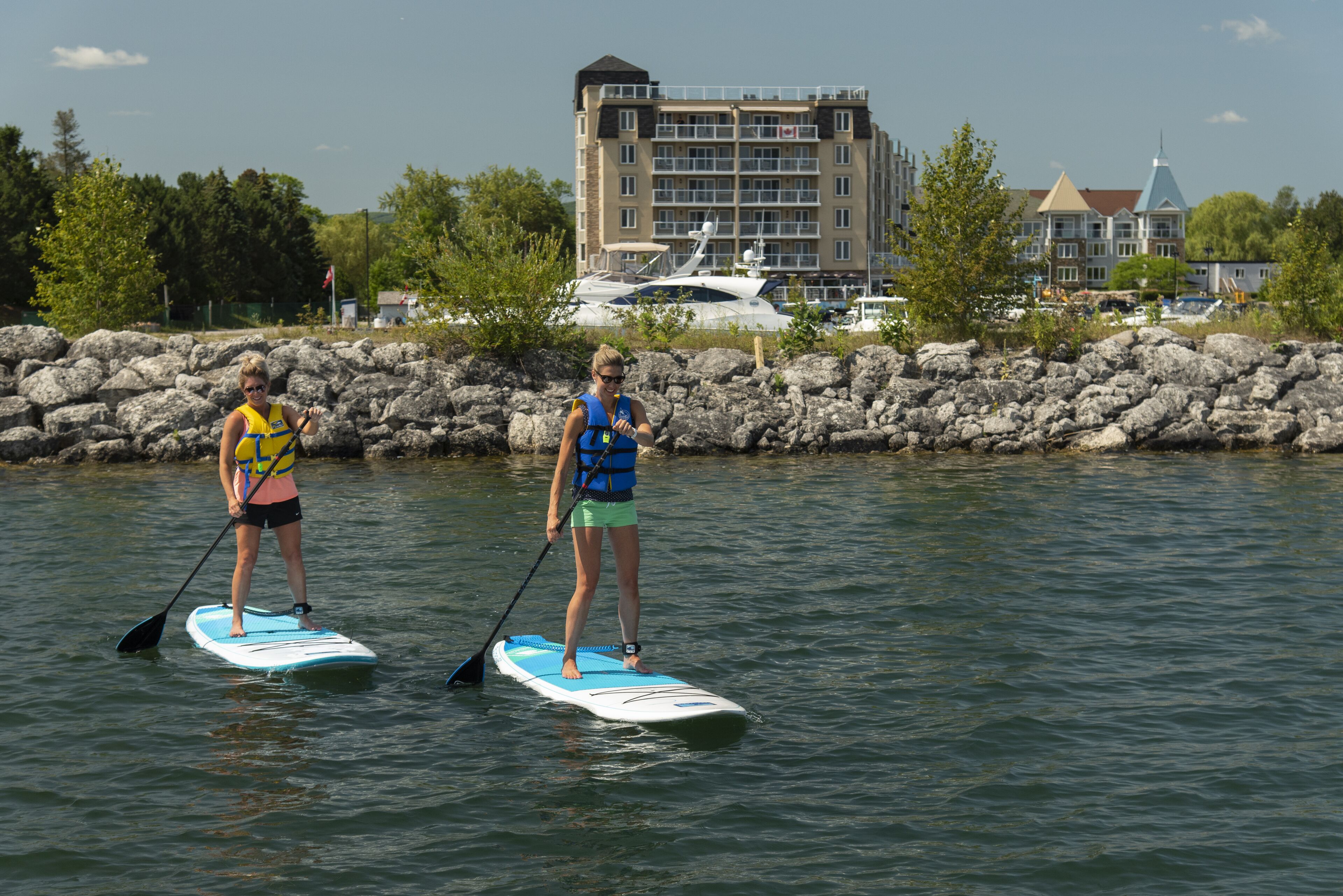 Two people paddleboarding on a calm body of water with a rocky shoreline and buildings in the background.