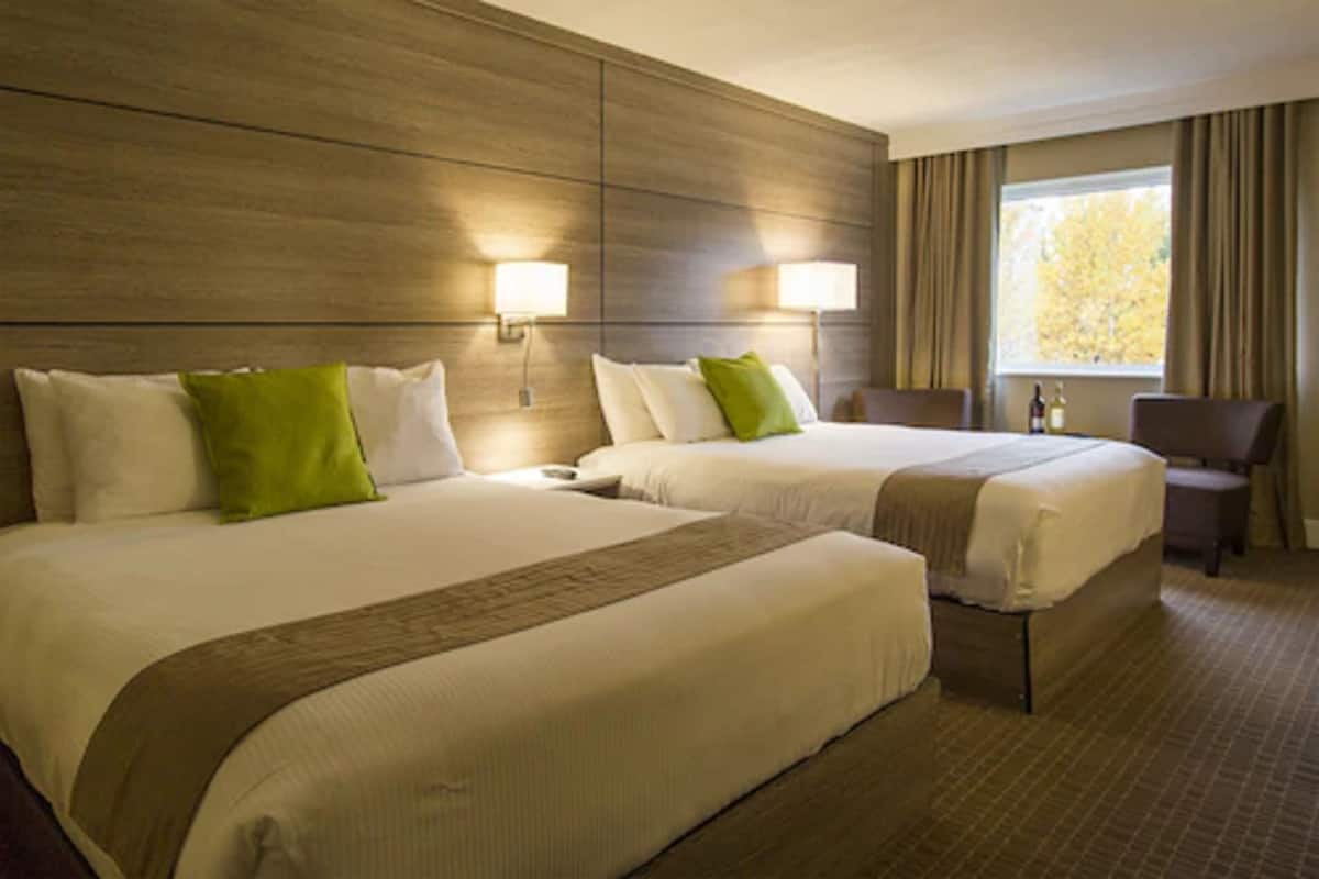 A hotel room with two beds, a desk, and a chair. There are two wall-mounted lamps and a window with curtains.