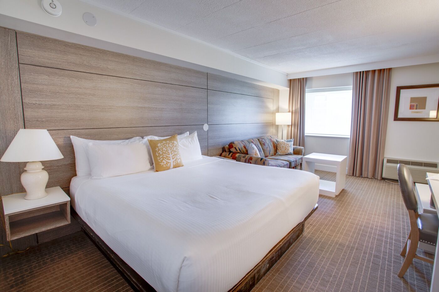 A hotel room with a large bed, a desk, a chair, a nightstand, and a window with curtains.