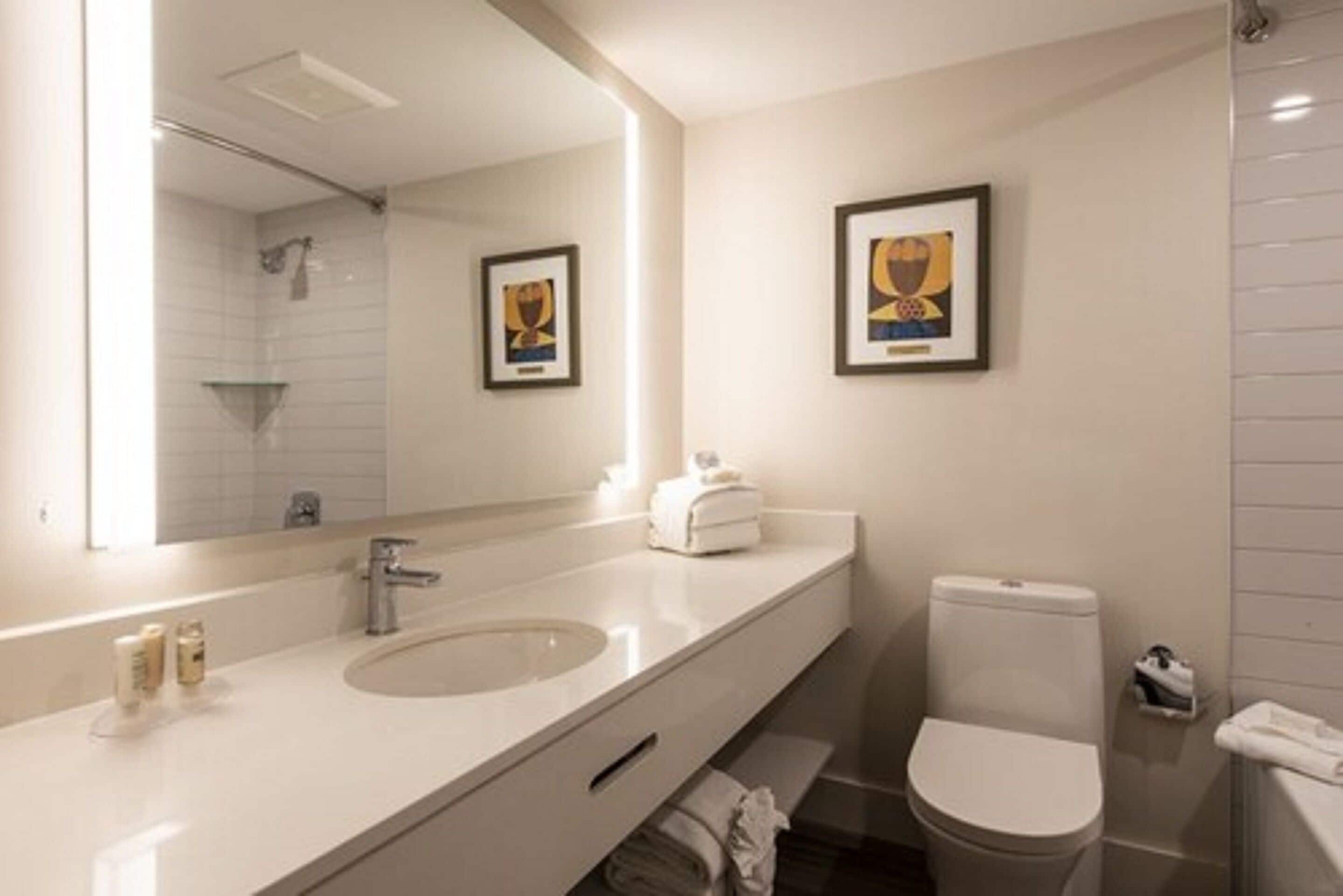 Un baño moderno con un espejo grande, dos lavamanos, un inodoro y obras de arte enmarcadas en la pared.