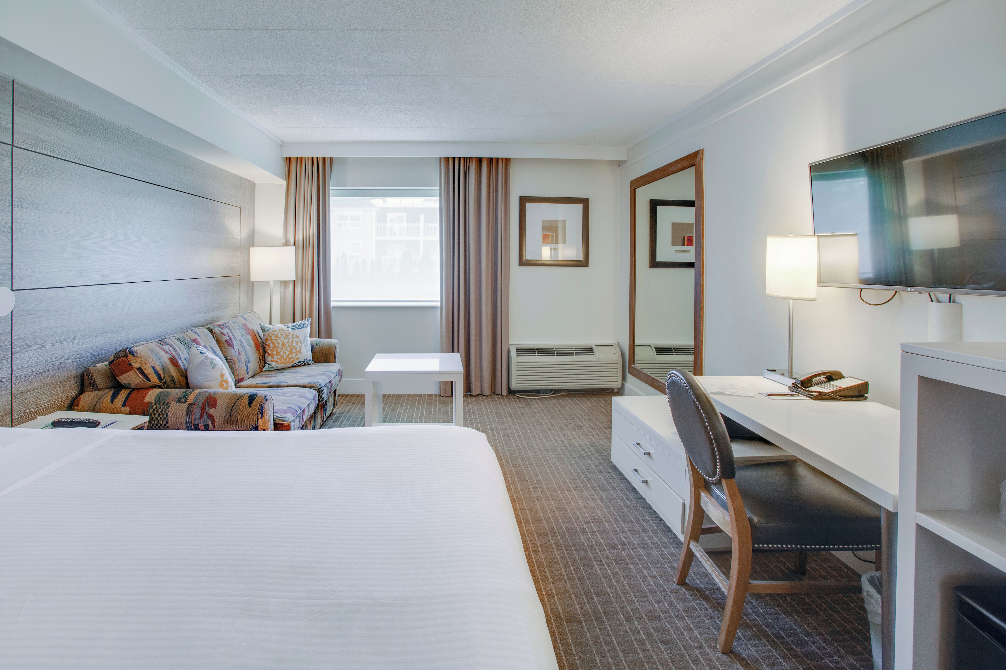 A hotel room with a large bed, a desk, a chair, a TV, a lamp, and a window with curtains.