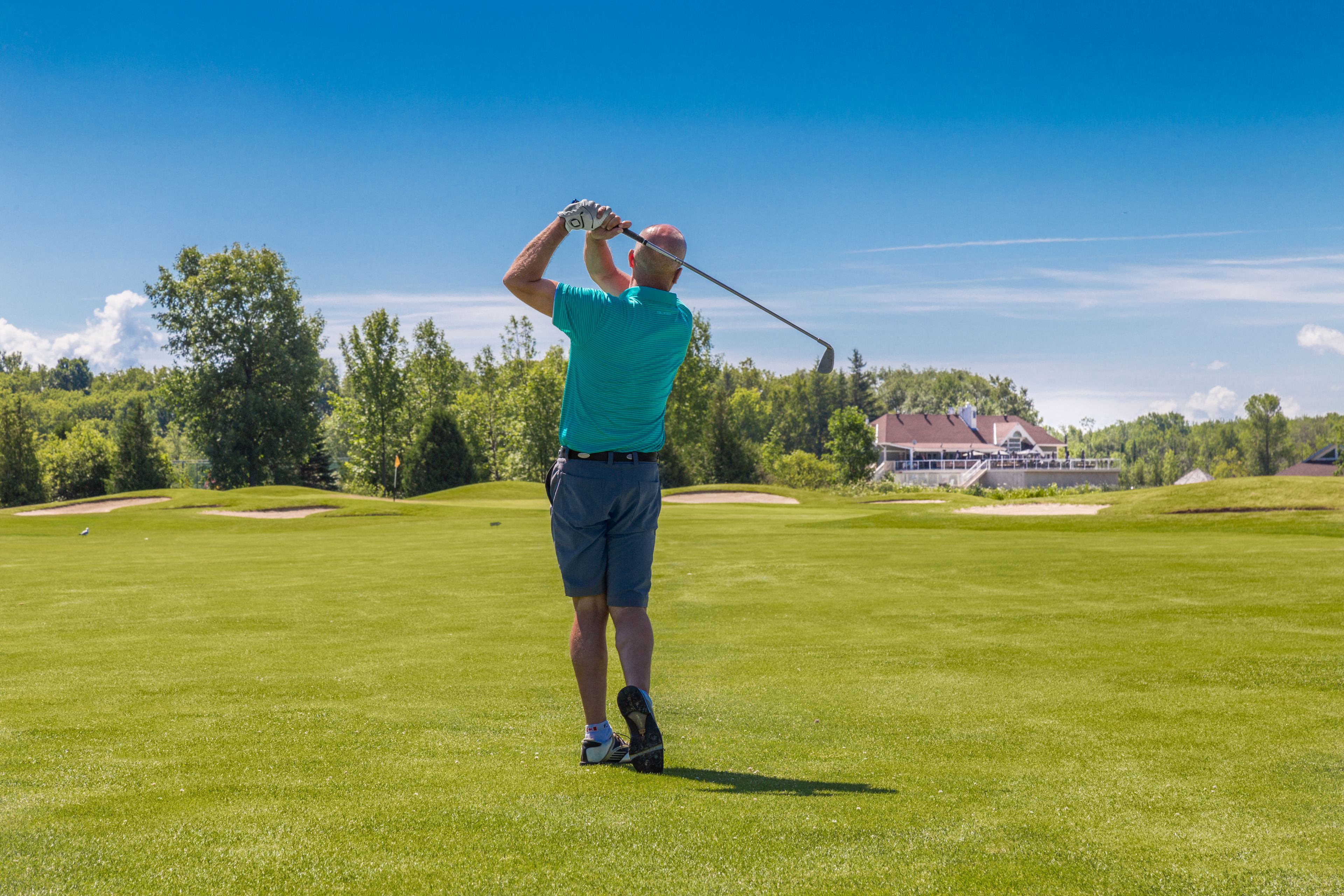 A golf course with a clear sky, a golfer in a teal shirt and shorts, and a building in the background.