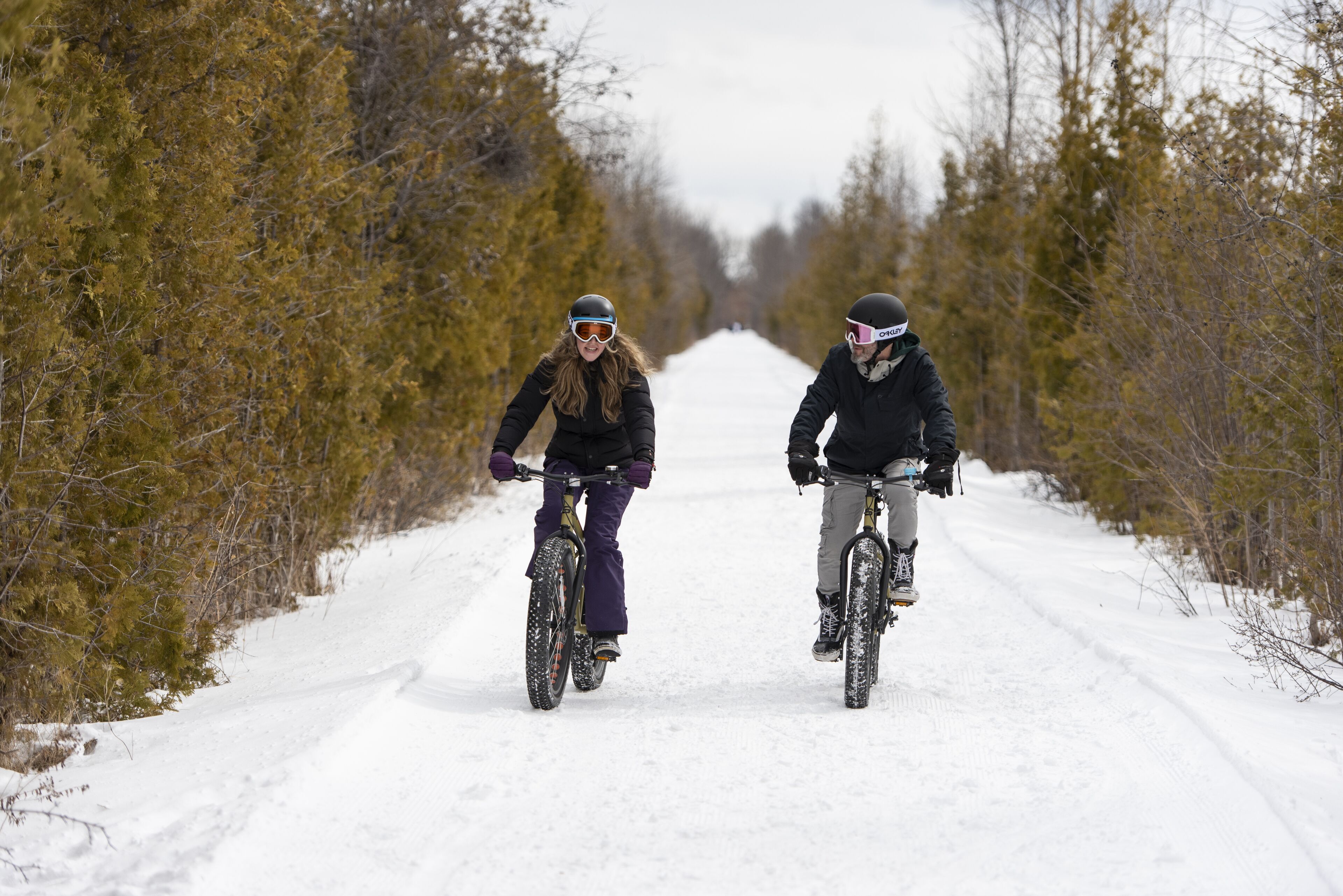 Dos personas paseando en bicicleta por un sendero cubierto de nieve, rodeado de árboles.