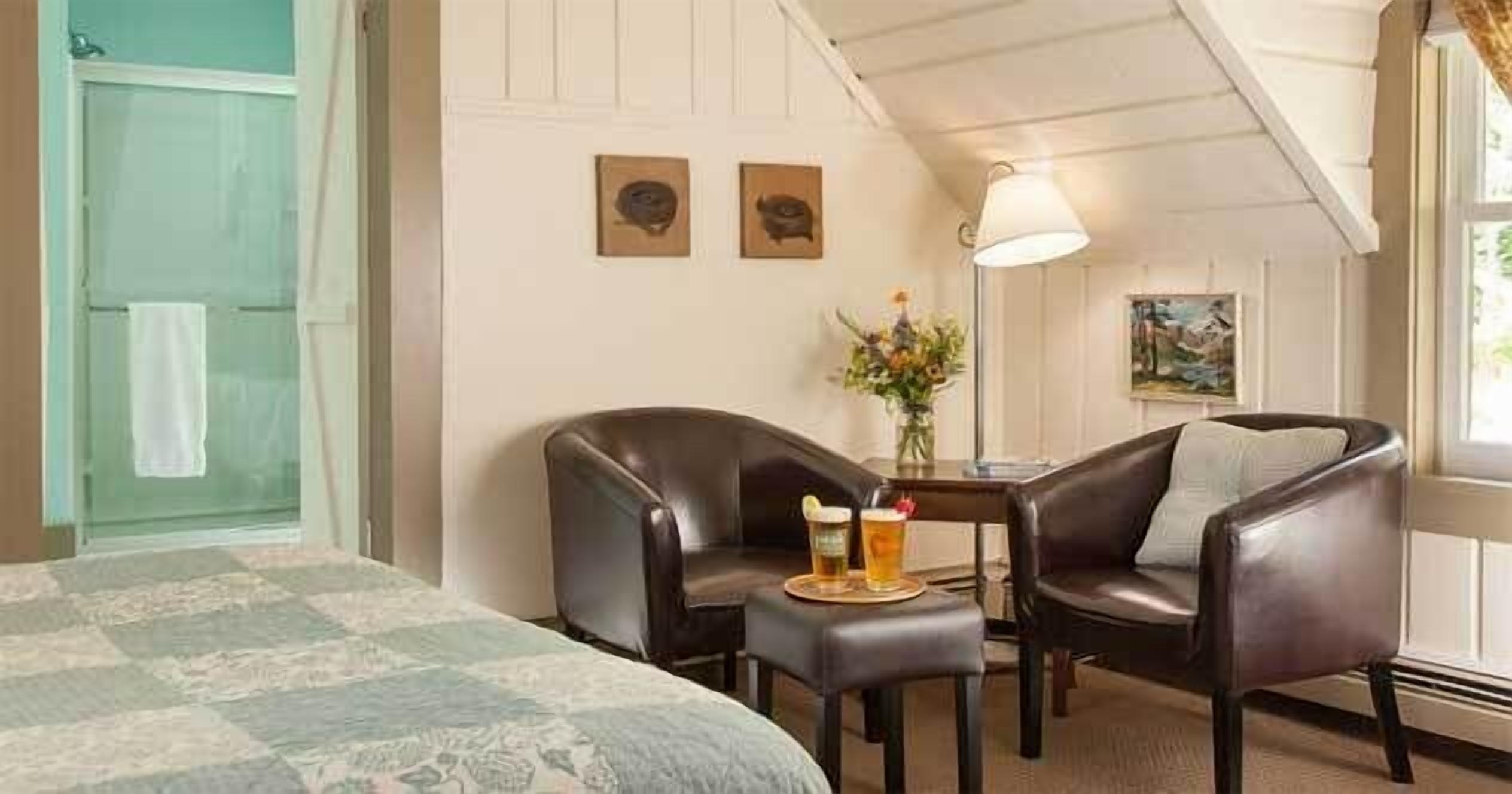 A hotel room with a bed, two leather chairs, a small table with a drink, and a painting on the wall.