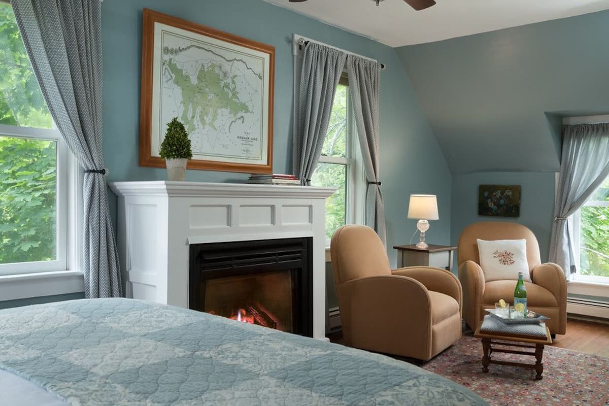Une chambre à coucher avec une cheminée, un lit recouvert d’une courtepointe, deux fauteuils, une petite table et une lampe.