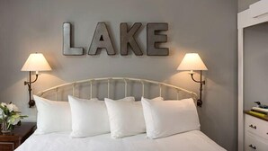 Una habitación de hotel con una cama, dos lámparas de noche, un cartel en la pared que dice 'LAKE' y una mesita de noche con un jarrón de flores.