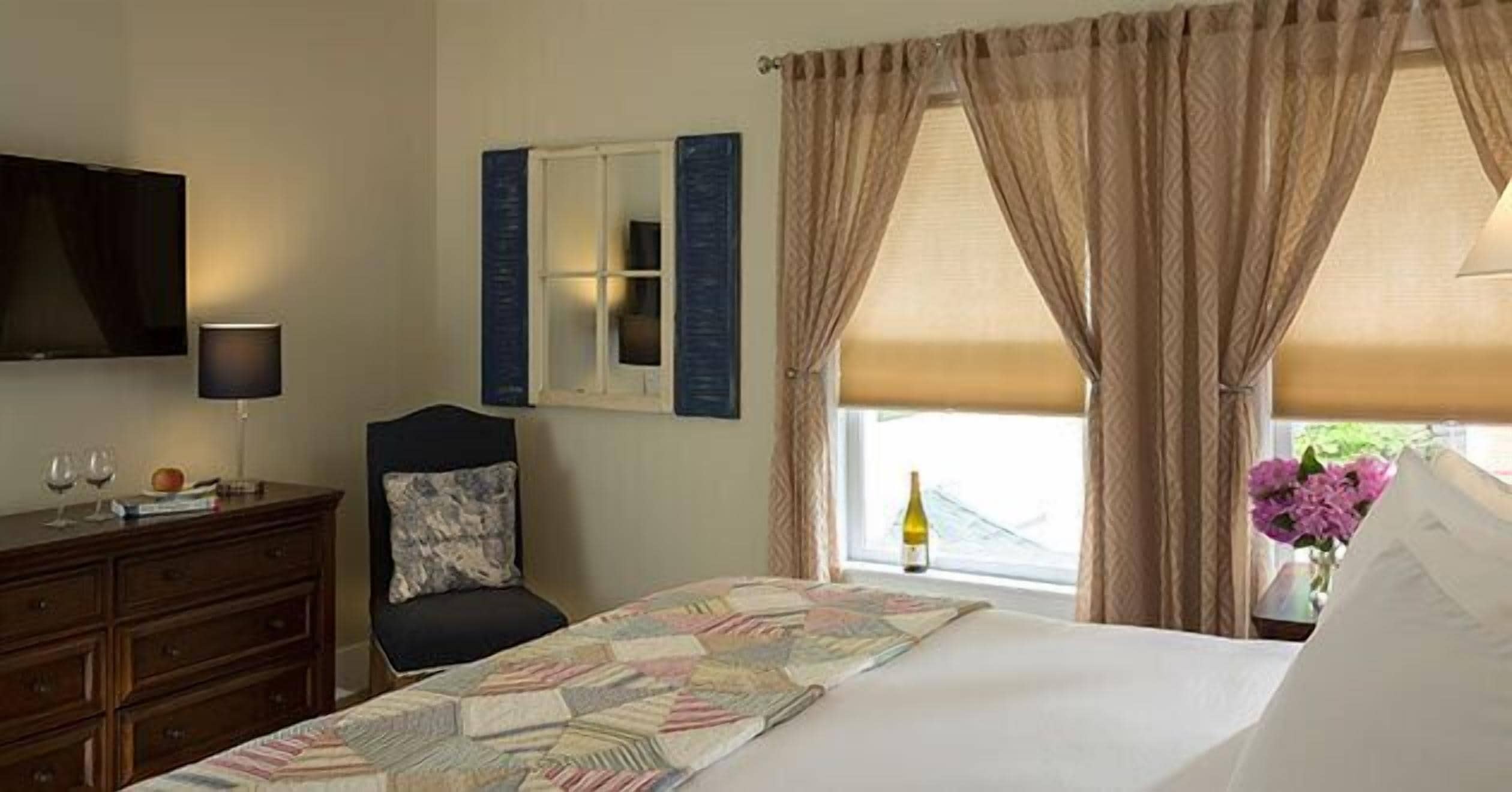 A hotel room with a bed, a dresser, a chair, a television, a window with curtains, and a vase with flowers.