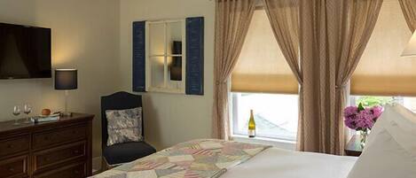 A hotel room with a bed, a dresser, a chair, a television, a window with curtains, and a vase with flowers.