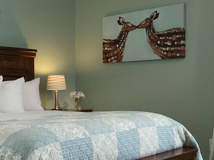 A bedroom with a bed, pillows, a nightstand with a lamp, a wall sign, and a painting.
