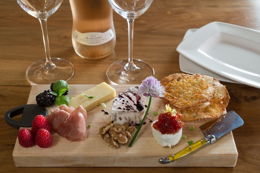 Une planche en bois garnie d’une variété de fromages, de fruits et de noix, accompagnée de deux verres à vin et d’une bouteille de vin.