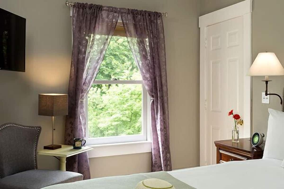 Une chambre d’hôtel comprenant un lit, une chaise, un bureau, une lampe, une fenêtre avec des rideaux et une lampe fixée au mur.