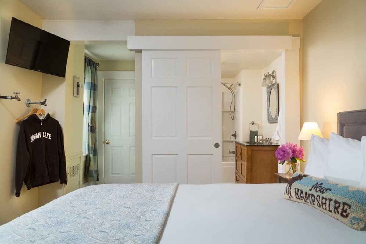 Une chambre d’hôtel avec un lit, un téléviseur fixé au mur, un porte-manteau avec une veste, une porte donnant sur une salle de bain, et une table de chevet avec une lampe et des fleurs.