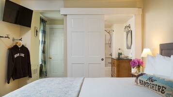 A hotel room with a bed, a TV mounted on the wall, a coat rack with a jacket, a door leading to a bathroom, and a bedside table with a lamp and flowers.