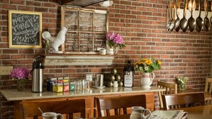 Una cocina rústica con un menú en una pizarra, una decoración de gallo blanco y una pared de ladrillo.