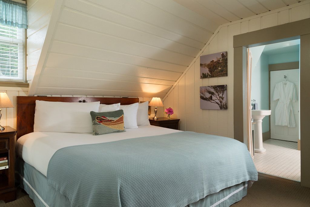 Un dormitorio con una cama grande, dos mesitas de noche con lámparas, una ventana con persianas y un baño visible a través de una puerta abierta.