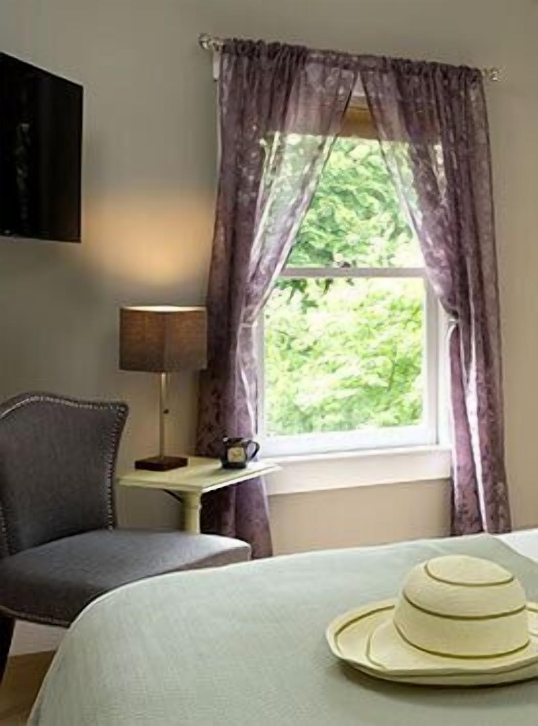 A hotel room with a bed, a chair, a small table with a lamp and a cup, and a window with curtains.