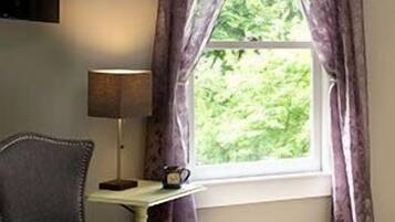 A hotel room with a bed, a chair, a small table with a lamp and a cup, and a window with curtains.