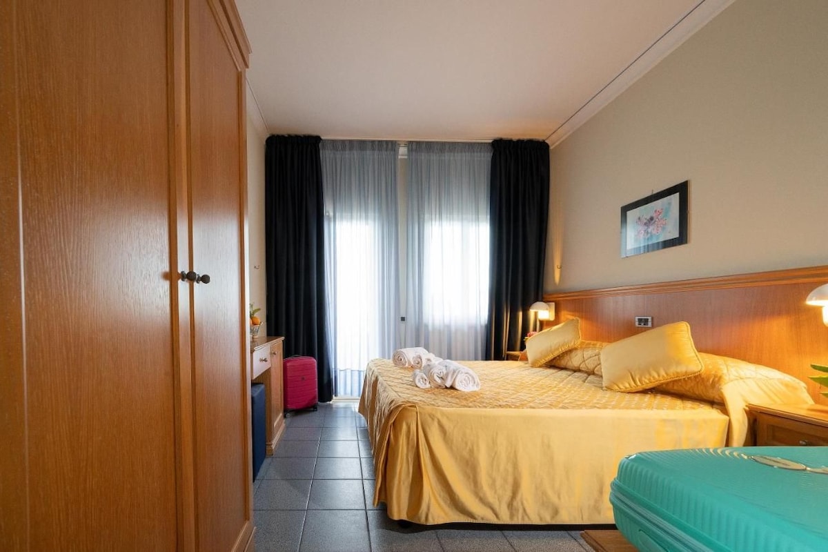 A hotel room with a bed, a nightstand, a lamp, a suitcase, and a wardrobe.