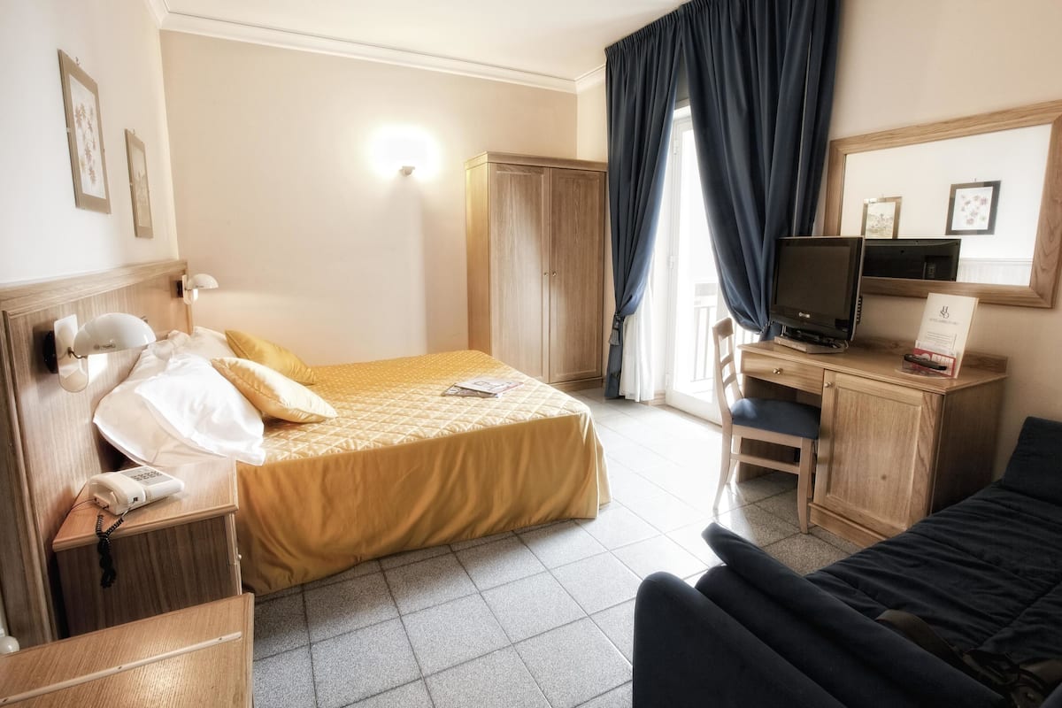 A hotel room with a bed, a desk with a computer, a chair, a wardrobe, and a TV.