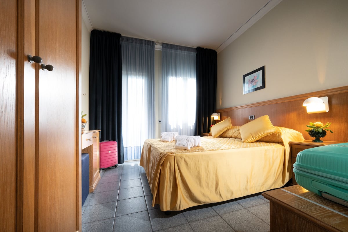 A hotel room with a bed, a suitcase, a nightstand with a lamp, and a window with curtains.