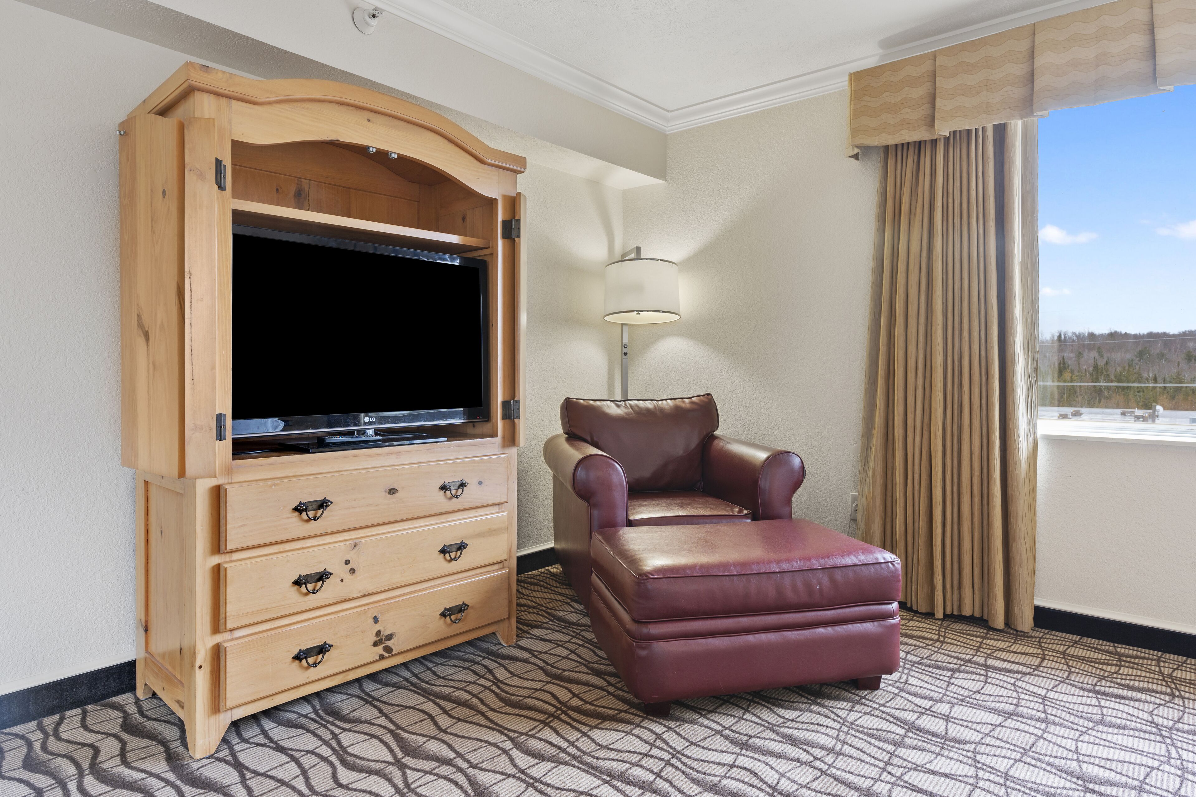 Habitación de hotel con una unidad de entretenimiento de madera, un sillón de cuero, una lámpara y vistas al exterior.