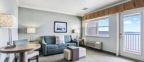 A modern living room with a blue sofa, a round table, and a small ottoman. There is a framed picture on the wall, a window with a view, and a door with a view.