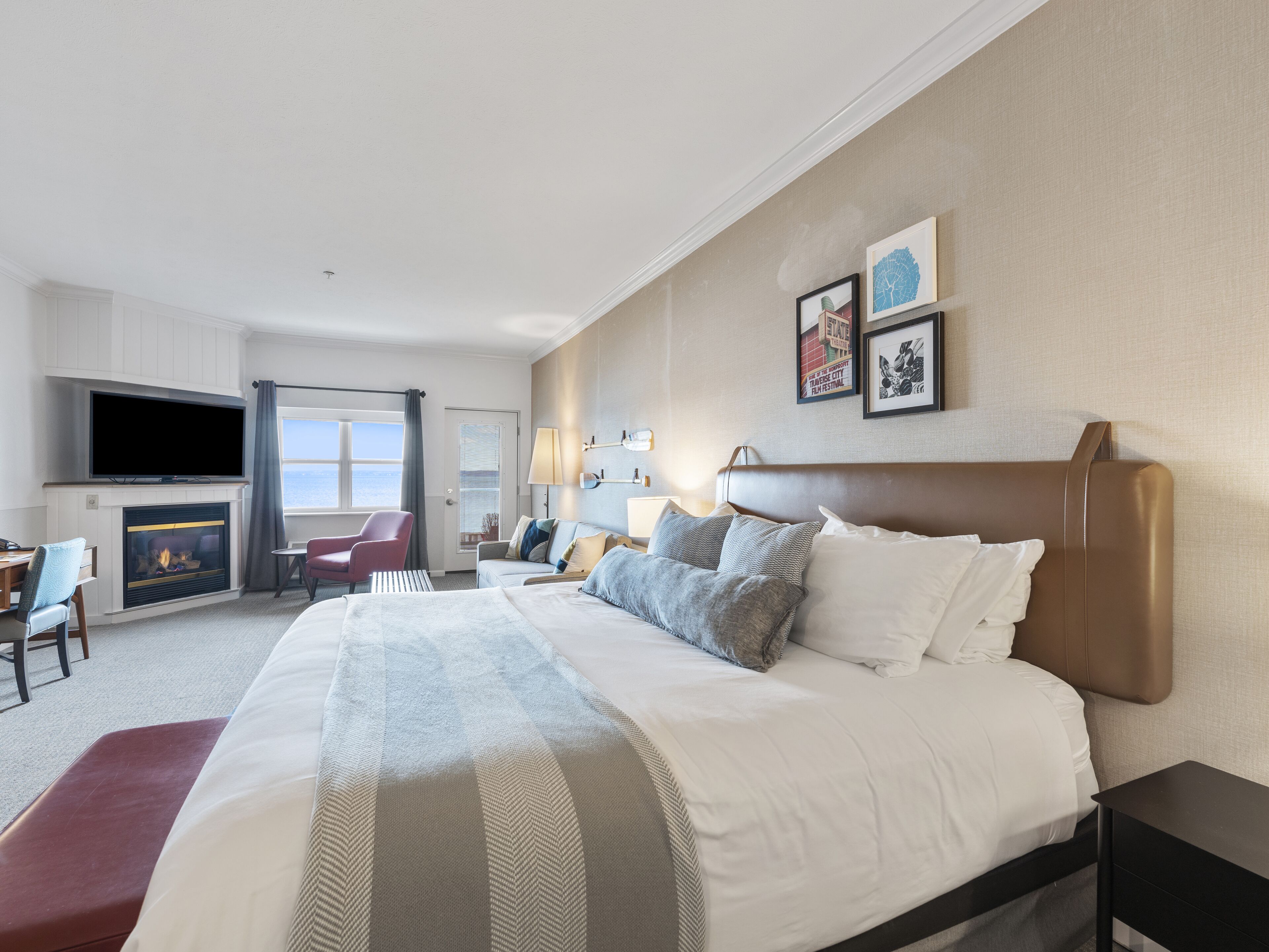 A hotel room with a large bed, a flat-screen TV mounted above a fireplace, a desk with a chair, and a view of the sea through a window.