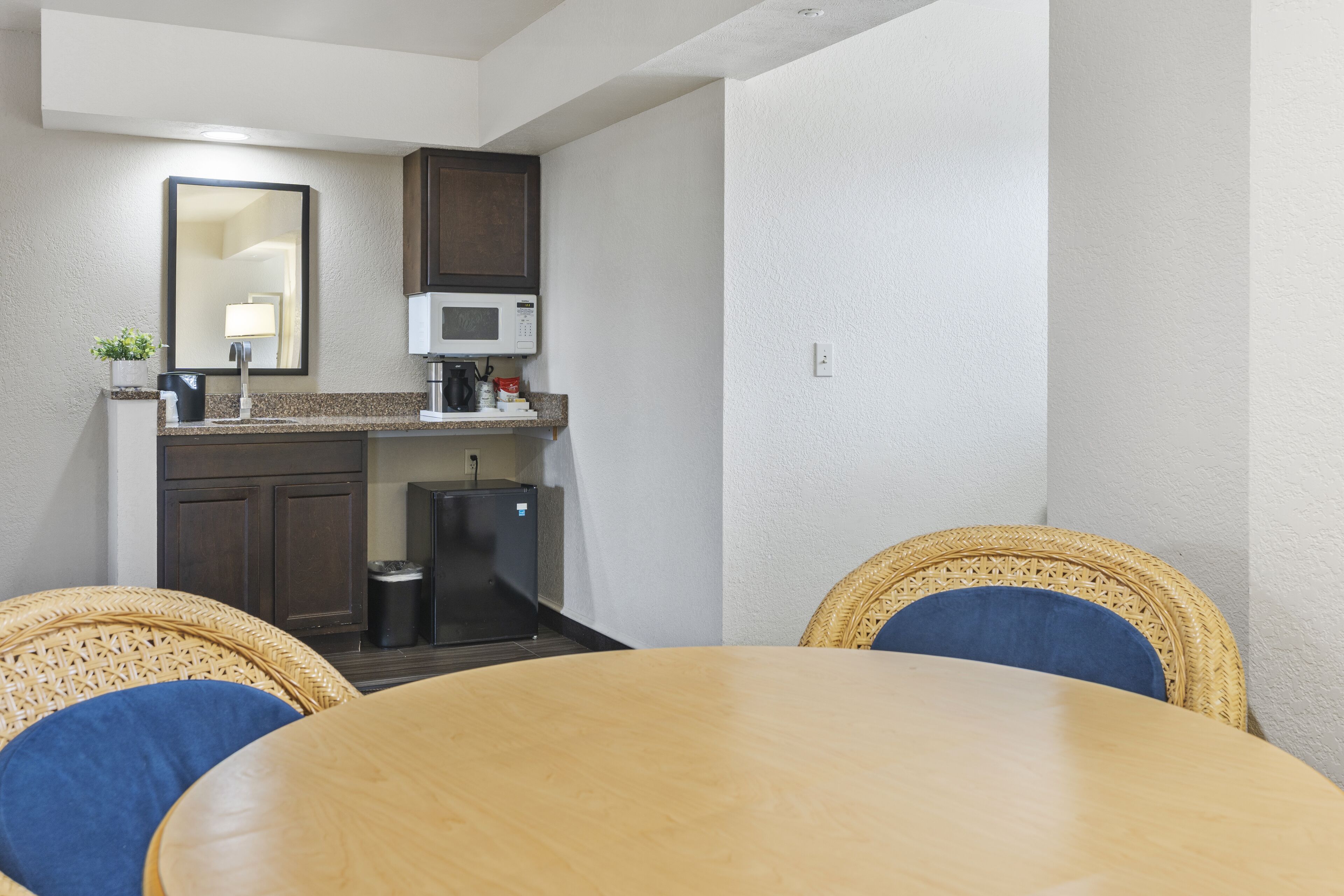 A compact kitchen with a microwave, a small refrigerator, and a sink. There are wicker chairs around a wooden table.