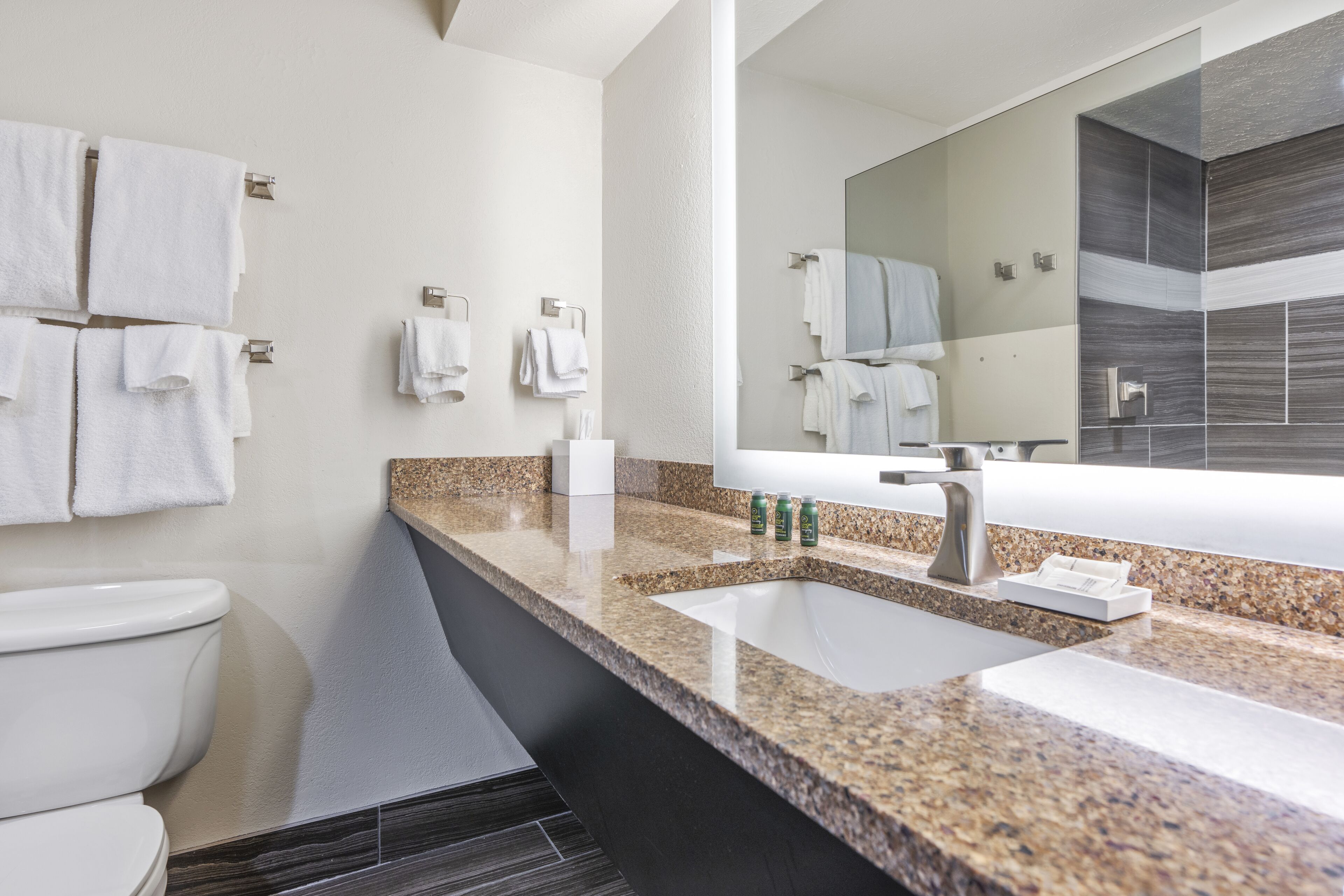 A modern bathroom with a granite countertop, two sinks, a large mirror, and multiple towel racks.