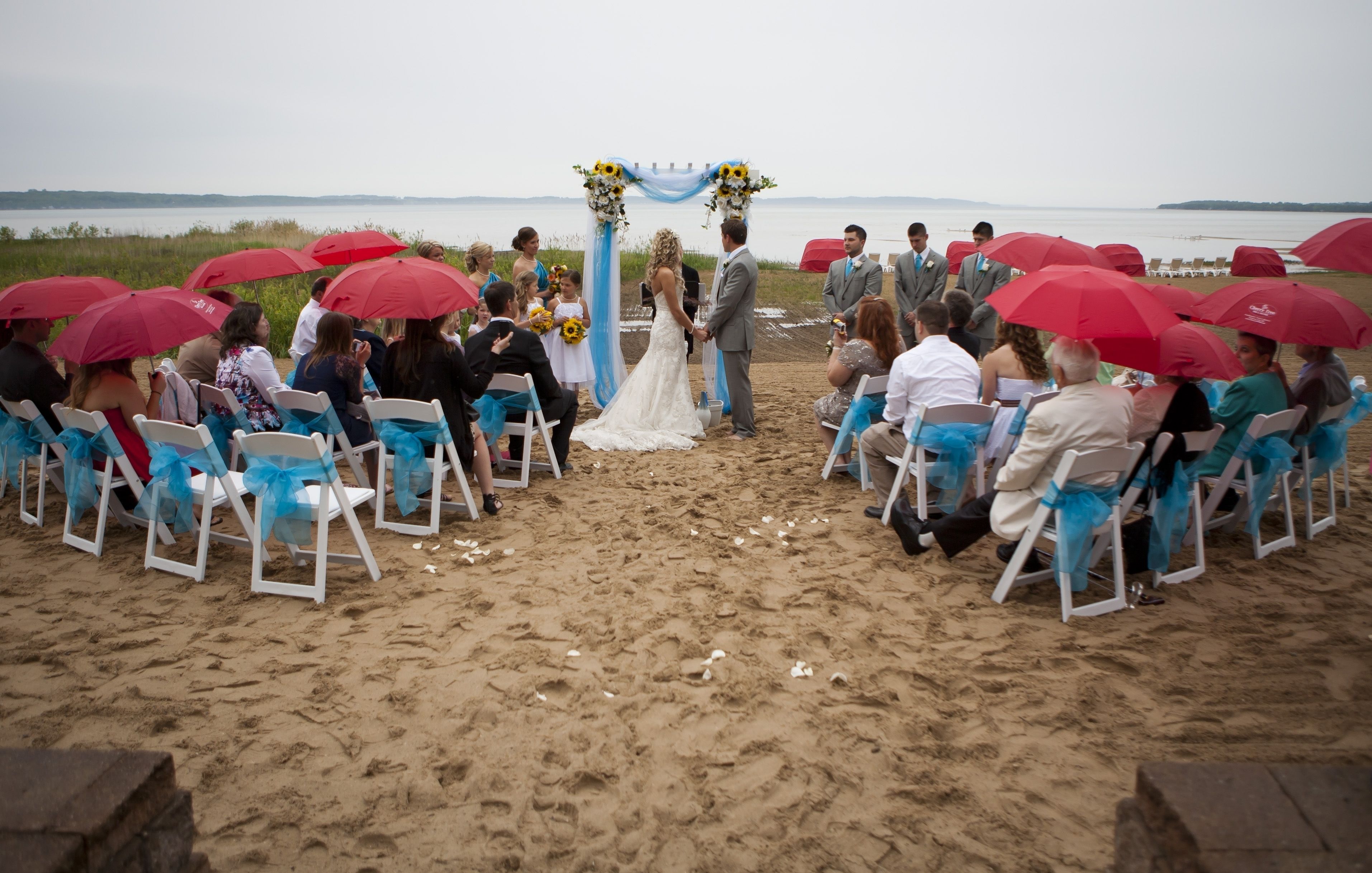 A beach wedding ceremony with guests seated on white chairs with blue sashes, a bride and groom standing under a floral arch, and red umbrellas providing shade.