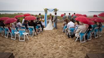 A beach wedding ceremony with guests seated on white chairs with blue sashes, a bride and groom standing under a floral arch, and red umbrellas providing shade.