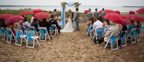 Cerimônia de casamento na praia com convidados sentados em cadeiras brancas com fitas azuis, um casal de noivos sob um arco floral, e guarda-sóis vermelhos oferecendo sombra.