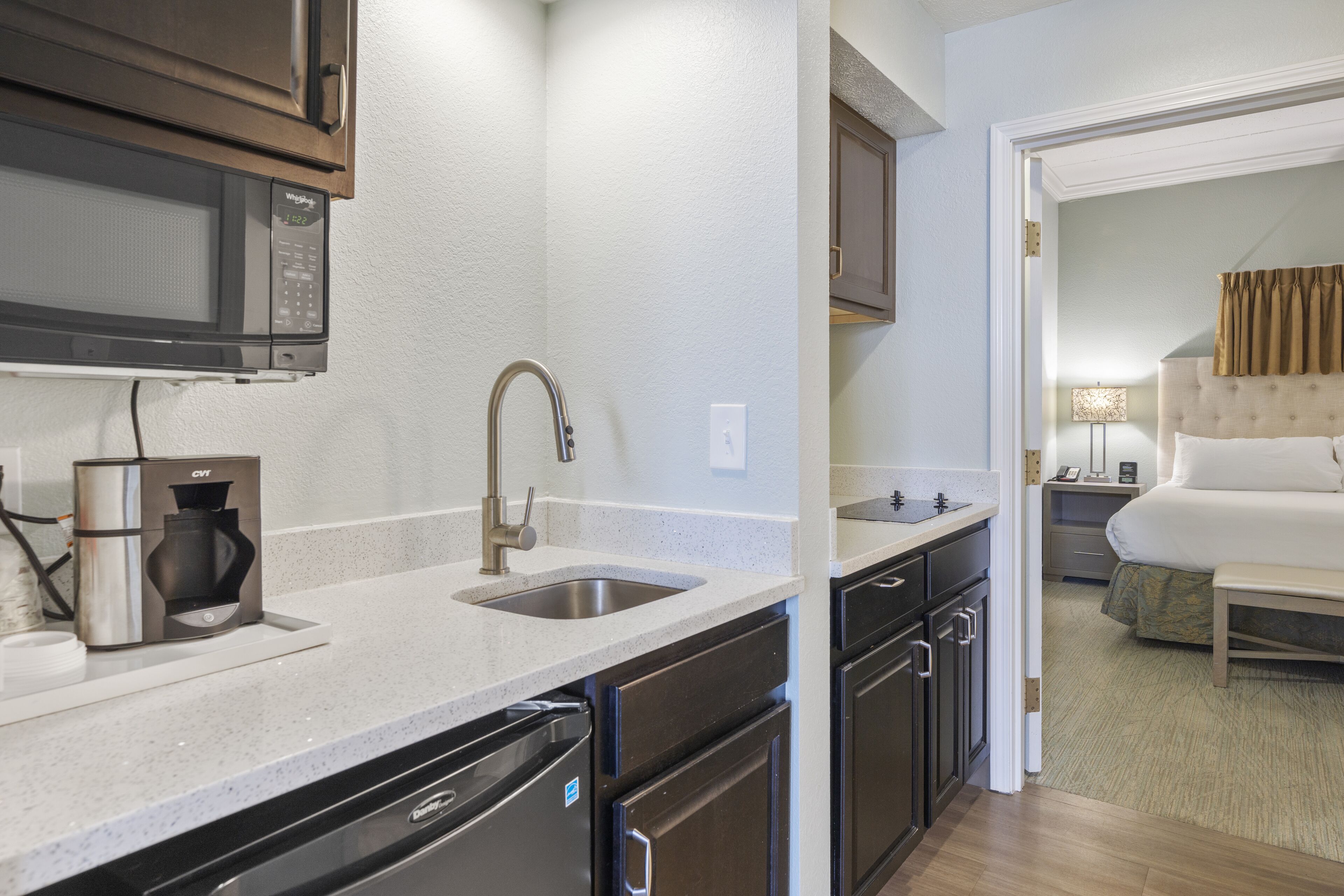 A compact kitchen with a microwave, coffee maker, sink, and dishwasher. A bedroom with a bed and nightstand is visible through an open doorway.