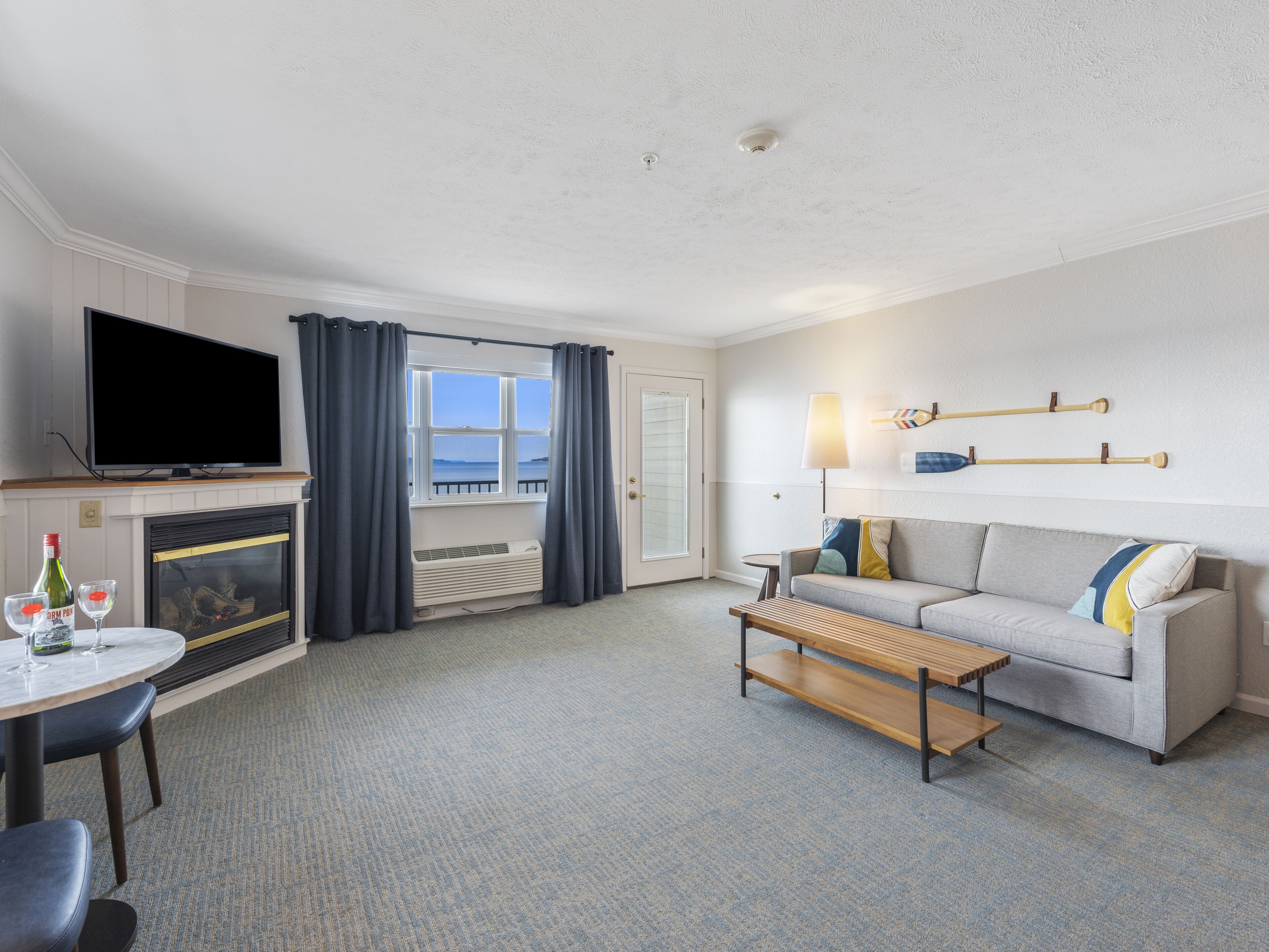 A hotel room with a TV, a sofa, a coffee table, a fireplace, a window with curtains, and a lamp.