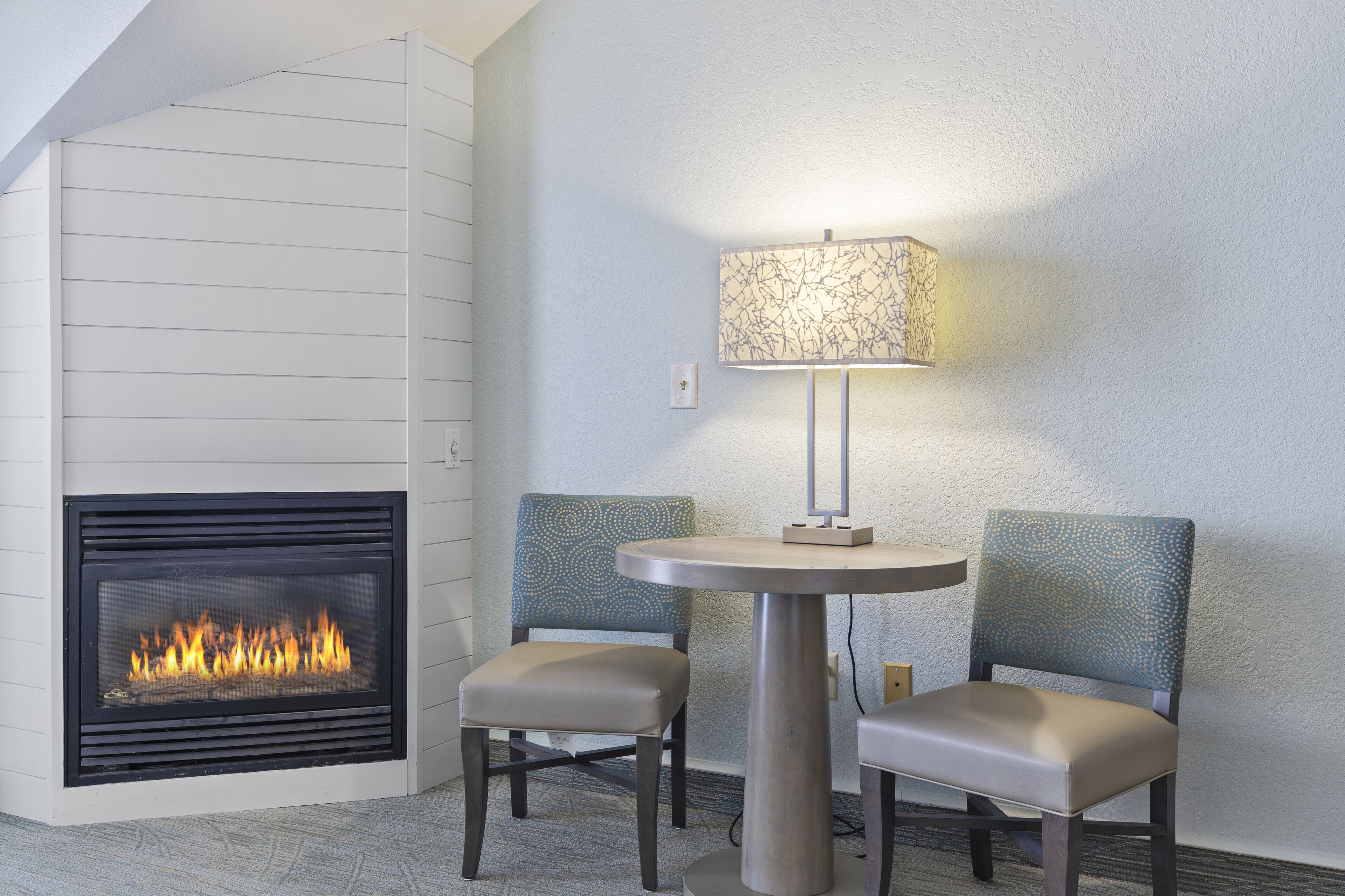 Een moderne open haard, twee gestoffeerde fauteuils en een tafel met een lamp.