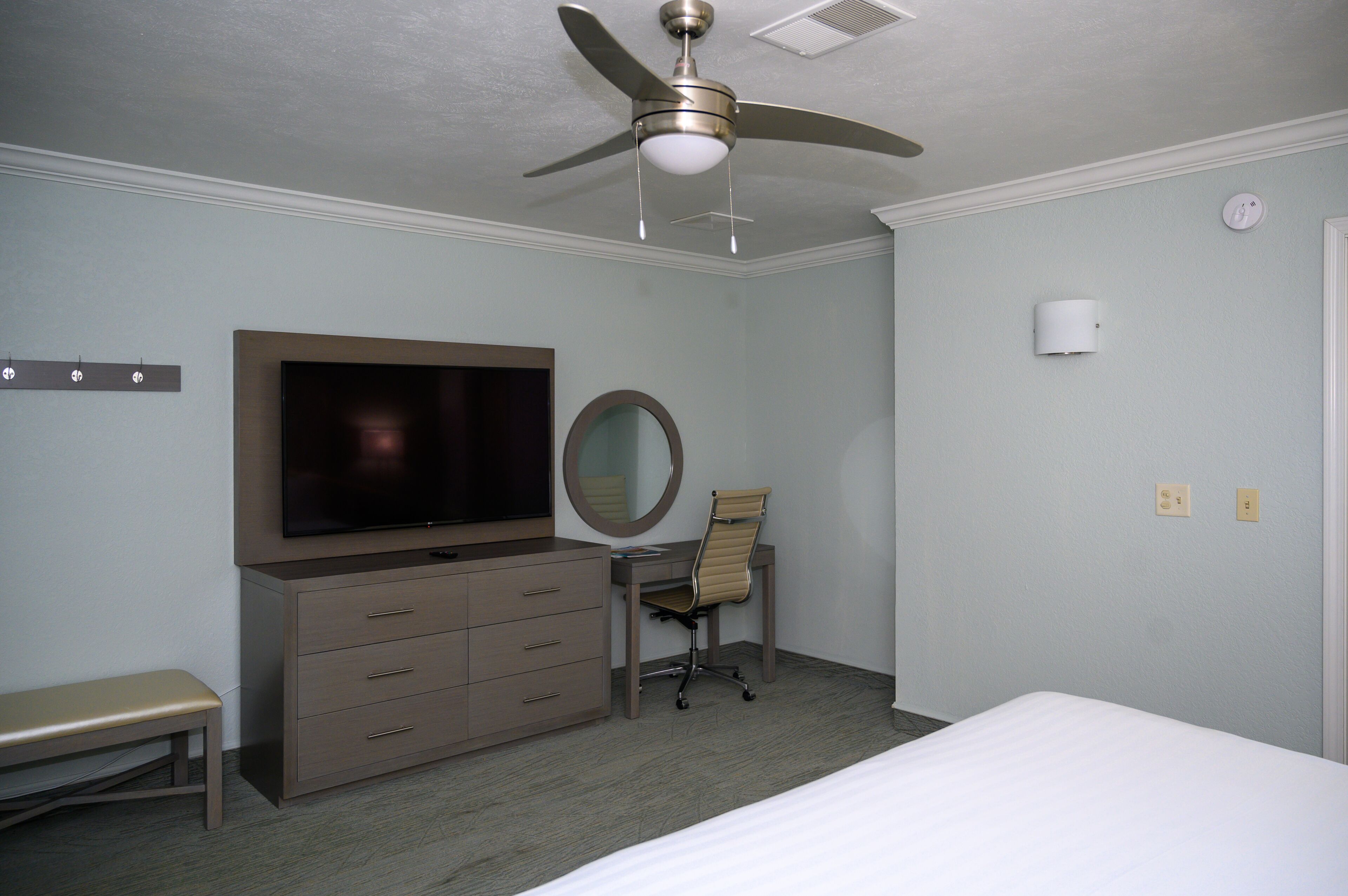 Quarto de hotel com uma cama, uma escrivaninha com cadeira, televisão, ventoinha de teto e espelho.