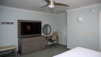 Quarto de hotel com uma cama, uma escrivaninha com cadeira, televisão, ventoinha de teto e espelho.