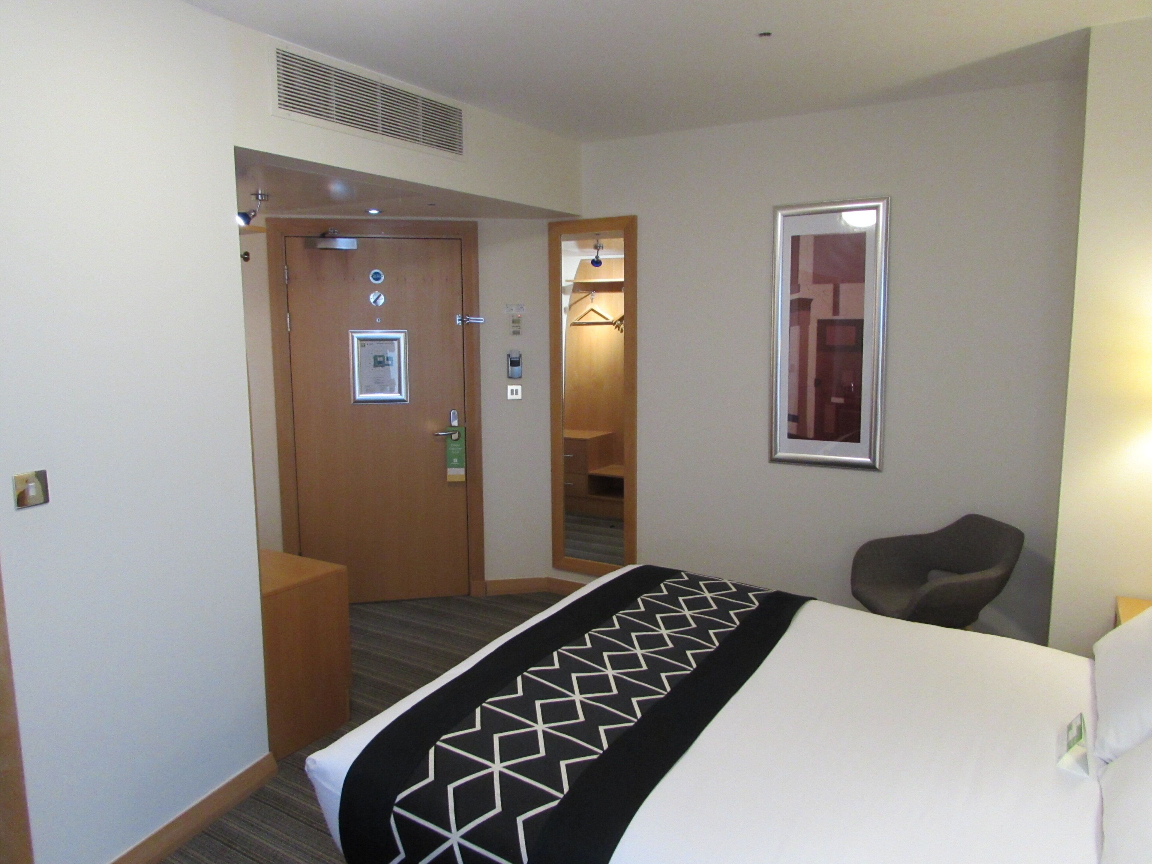 A hotel room with a bed, a chair, a door, a mirror, and a wall-mounted light fixture.