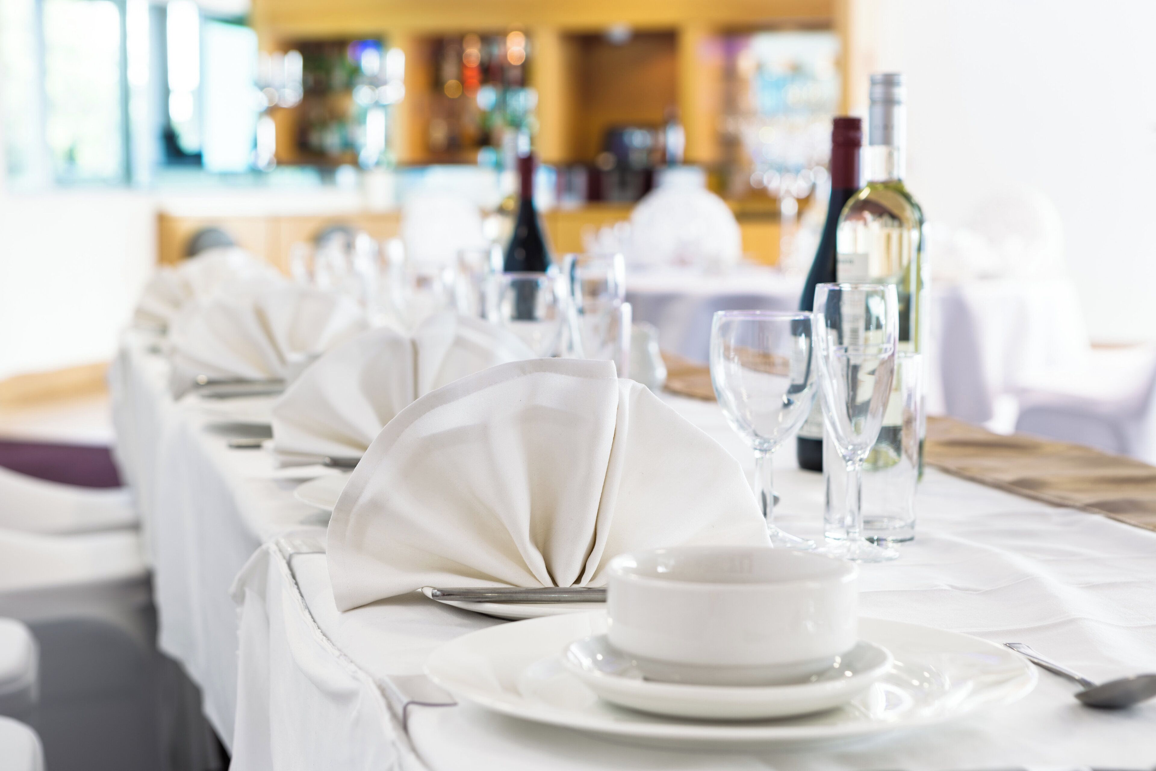 A dining table set with white tablecloths, plates, cutlery, and wine glasses, indicating a formal dining arrangement.