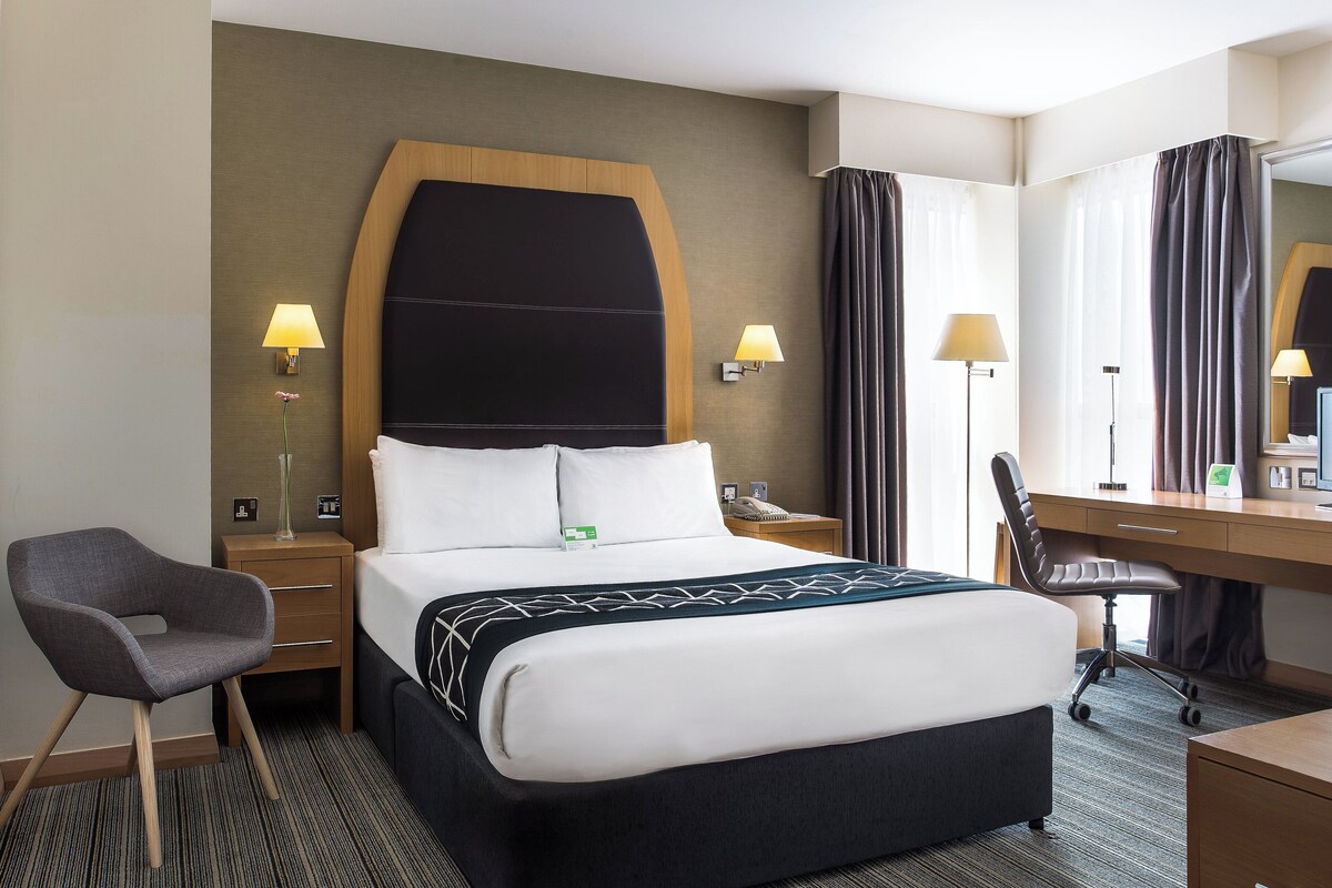 A hotel room with a bed, a desk, a chair, a lamp, and a mirror.