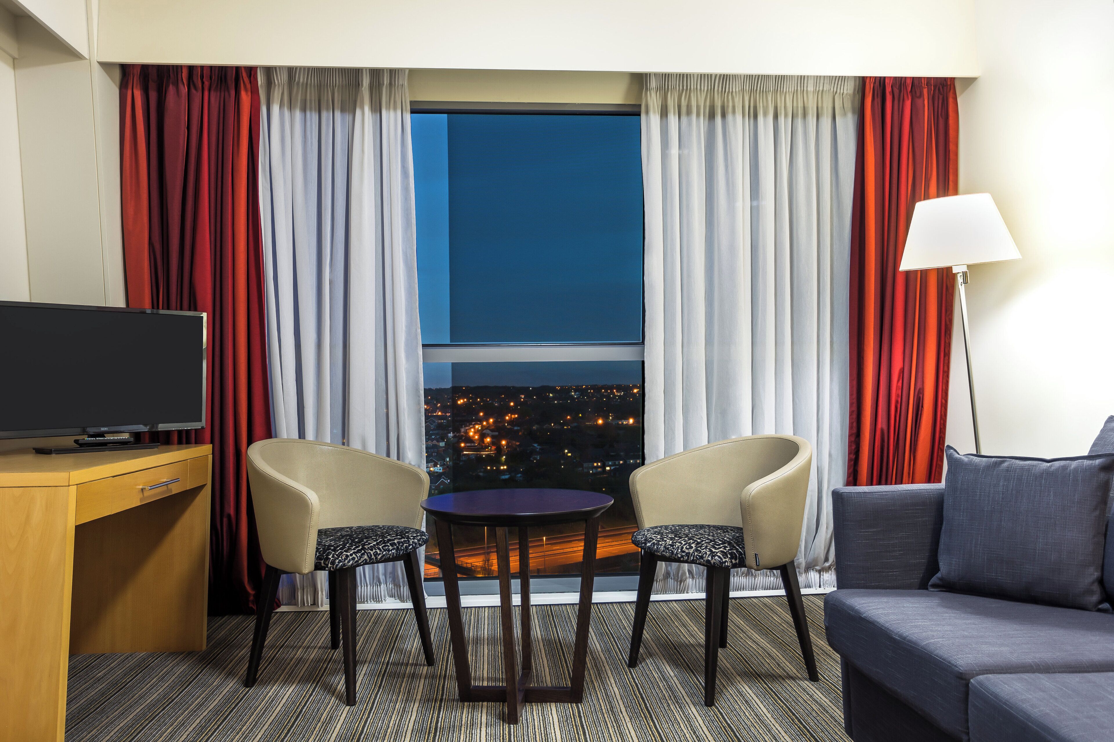 A hotel room with a television, two chairs, a small table, a sofa, and a view of a cityscape at night.