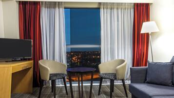 A hotel room with a television, two chairs, a small table, a sofa, and a view of a cityscape at night.