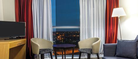 A hotel room with a television, two chairs, a small table, a sofa, and a view of a cityscape at night.